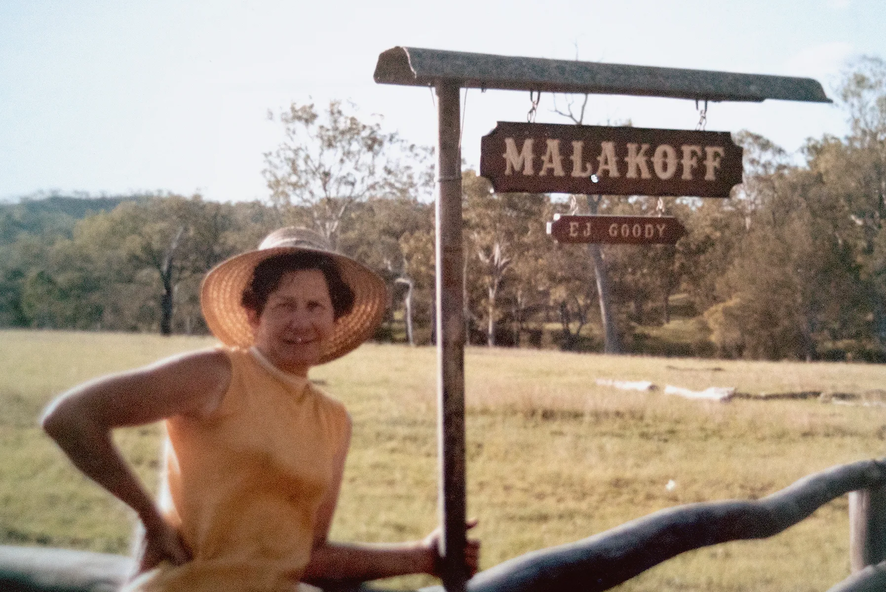 Girlie wears a yellow top adn a sunhat and leans against the sign at the front of her property, which reads, 'Malakoff, EJ Goodie.'