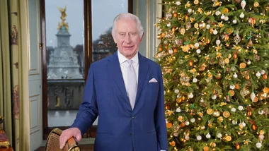 A man in a blue suit stands near a decorated Christmas tree indoor.
