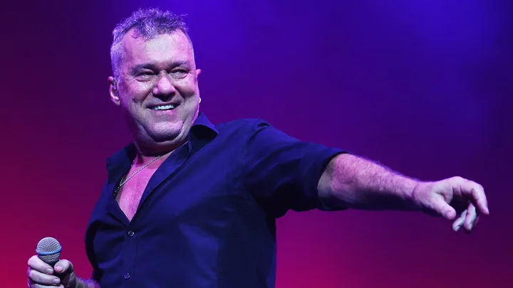 Man in a dark shirt holding a microphone on stage, pointing with his other hand, under purple lighting.