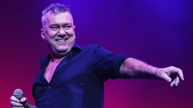Man in a dark shirt holding a microphone on stage, pointing with his other hand, under purple lighting.