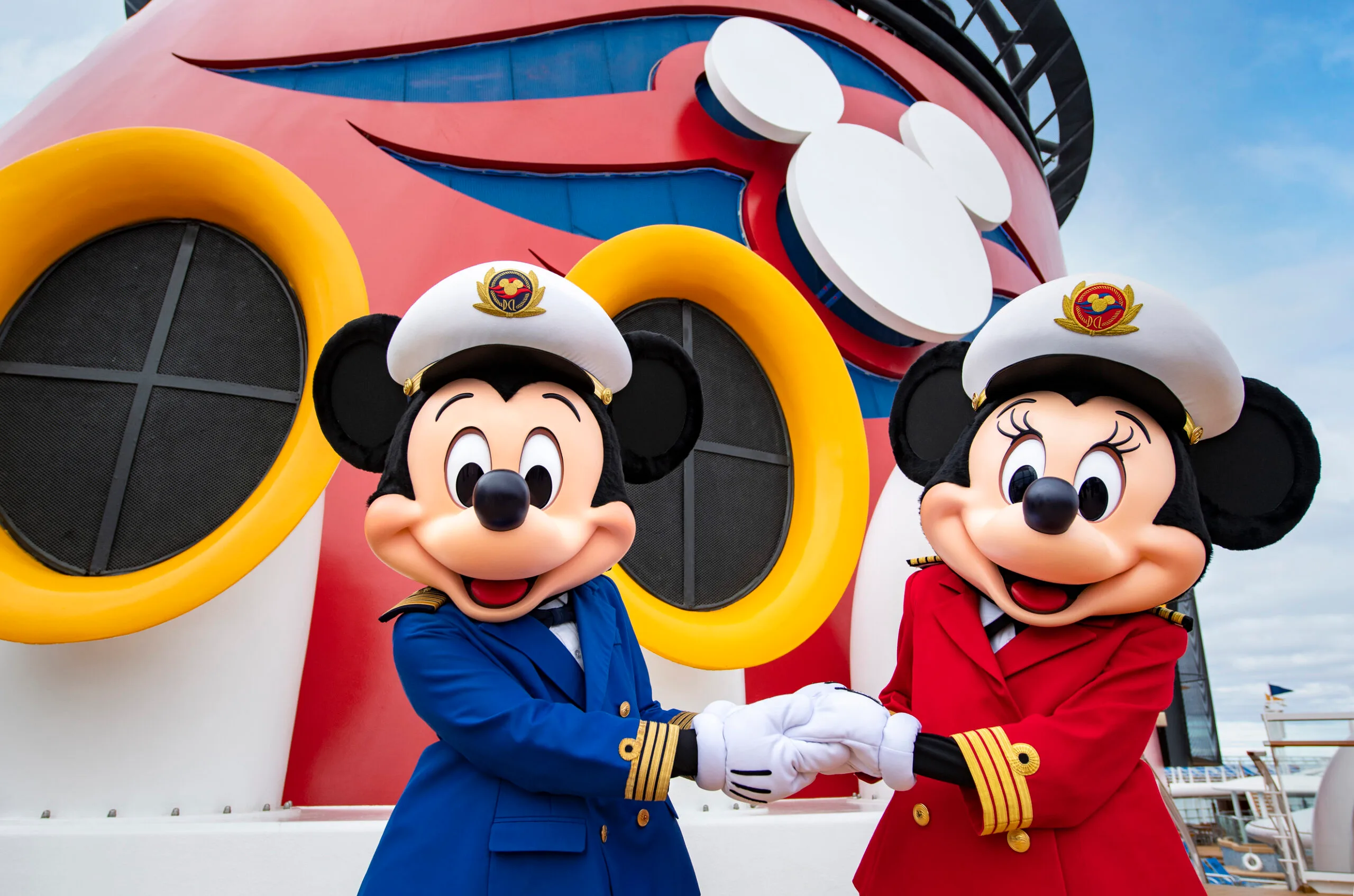 Mickey and Minnie Mouse in captain outfits, smiling and holding hands on a Disney Cruise ship.