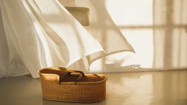 A wicker baby bassinet sits on a sunlit floor with a flowing white curtain in the background.