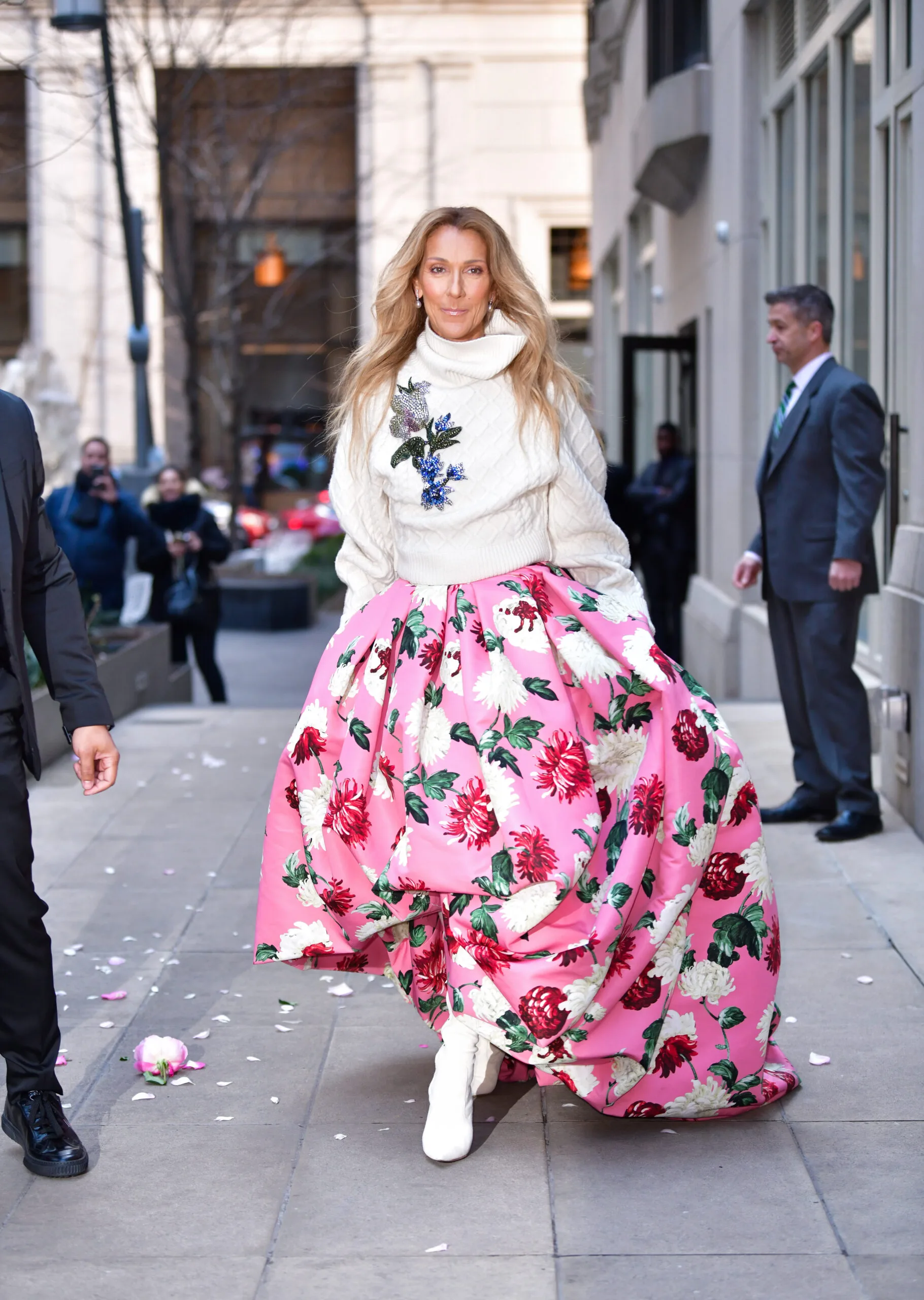 A woman in a floral pink skirt and white sweater walks confidently on a city street, accompanied by a man in a suit.