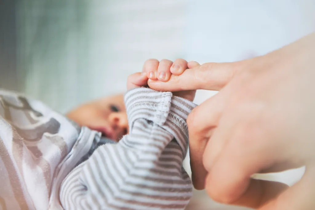 A baby in striped clothes holds an adult's finger, symbolizing connection and tenderness.