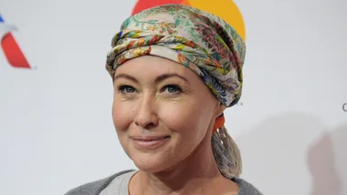 A person smiling, wearing a floral headscarf, with a neutral background.