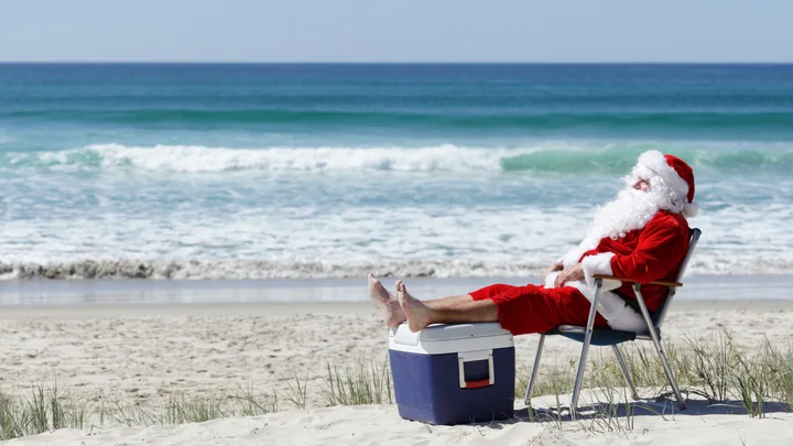 Santa Claus relaxing in a chair on a sandy beach, feet on a cooler, with ocean waves in the background.