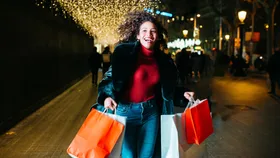 Smiling woman with curly hair carries shopping bags on a festive, illuminated street at night.