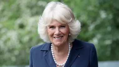 A woman with short blond hair wearing a navy suit and pearl necklace, smiling in front of a blurred green background.