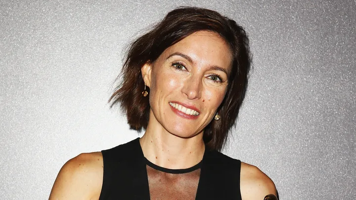 A woman with short brown hair and a black dress smiles against a glittery background.