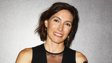 A woman with short brown hair and a black dress smiles against a glittery background.