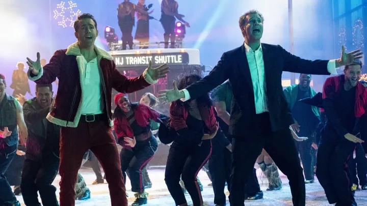 Two men wearing festive outfits lead a lively dance with a group in front of a bus labeled "NJ Transit Shuttle."