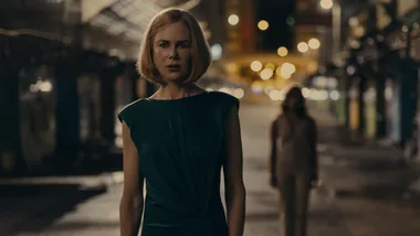 A woman in a green dress stands on a dimly lit street at night, with a blurred figure in the background.