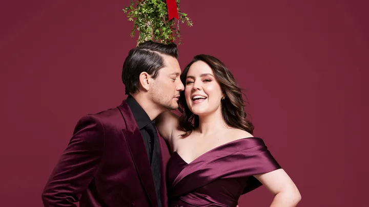 A couple in burgundy outfits stands closely under a mistletoe, against a matching burgundy background.