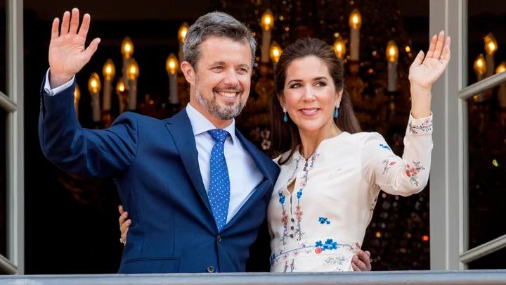 A man and a woman smiling and waving from a balcony with lit candles in the background.