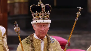 A man wearing a royal robe and ornate crown holds two scepters indoors.