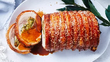 Roast pork with crispy crackling, slices revealing herb stuffing, on a white plate with bay leaves.