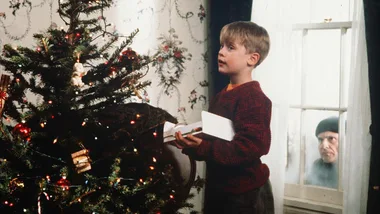 A young boy in front of a decorated Christmas tree, with a man peering through a window from outside in "Home Alone".