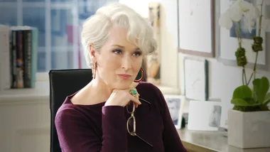 A woman with short white hair sits thoughtfully, holding glasses, in an office setting with books and white flowers.