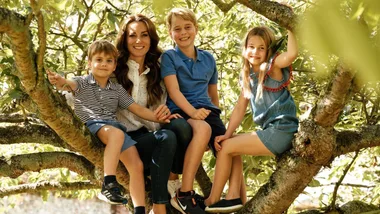 A woman and three children smile while sitting on tree branches with greenery around.
