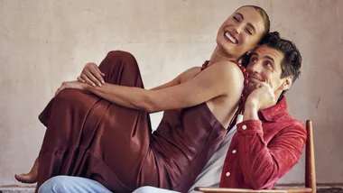 Man in red jacket seated with a smiling woman in a brown dress sitting on his lap, in a cozy setting.