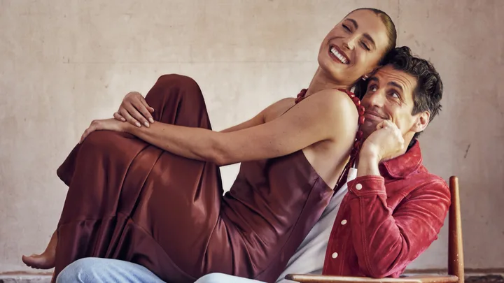 Man in red jacket seated with a smiling woman in a brown dress sitting on his lap, in a cozy setting.