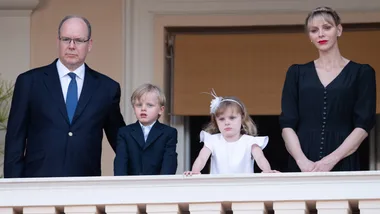 A family of four stands on a balcony. The adults wear formal attire, and the children wear a suit and a white dress.