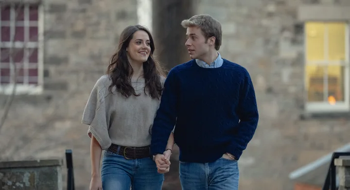 A couple holding hands and walking outdoors, both wearing casual sweaters and jeans, smiling at each other.