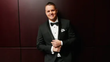 Man in a tuxedo smiling, standing against a dark wall.