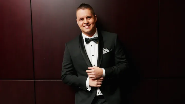 Man in a tuxedo smiling, standing against a dark wall.