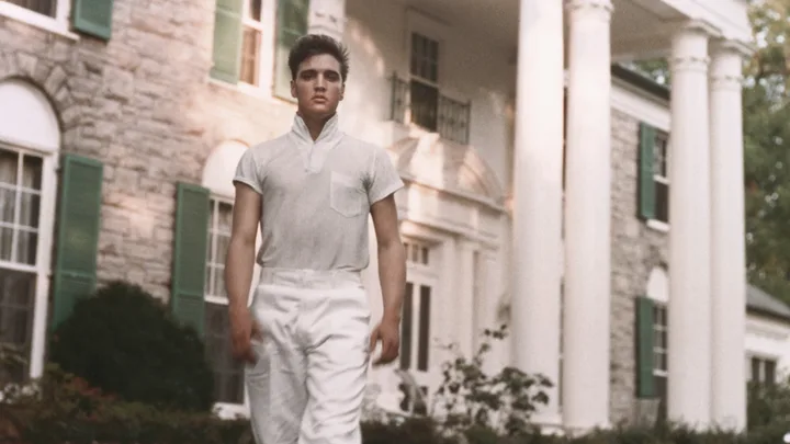 A person in a white outfit stands in front of a large stone house with green shutters and white columns.