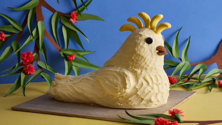Cockatoo-shaped cake with banana crest, chocolate eyes, and biscuit beak, set against colorful paper foliage.