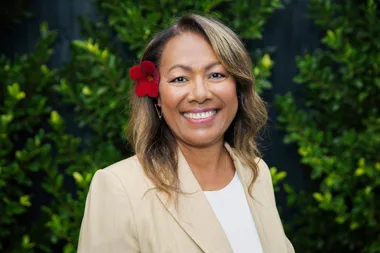A woman with a red flower in her hair smiles, standing in front of green foliage.