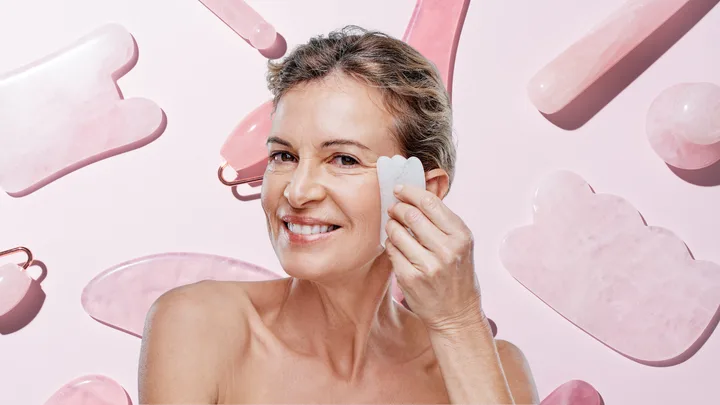 A woman smiling and using a pink gua sha tool on her face, with a pink background displaying more facial tools.