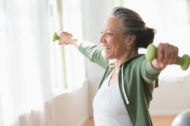 Elderly woman smiling while lifting small green dumbbells indoors, wearing a green hoodie and white top.