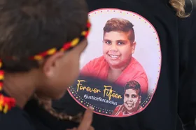 A child wearing a headband looks at a shirt with a photo of a smiling boy, text reads "Forever Fifteen #justiceforcassius."