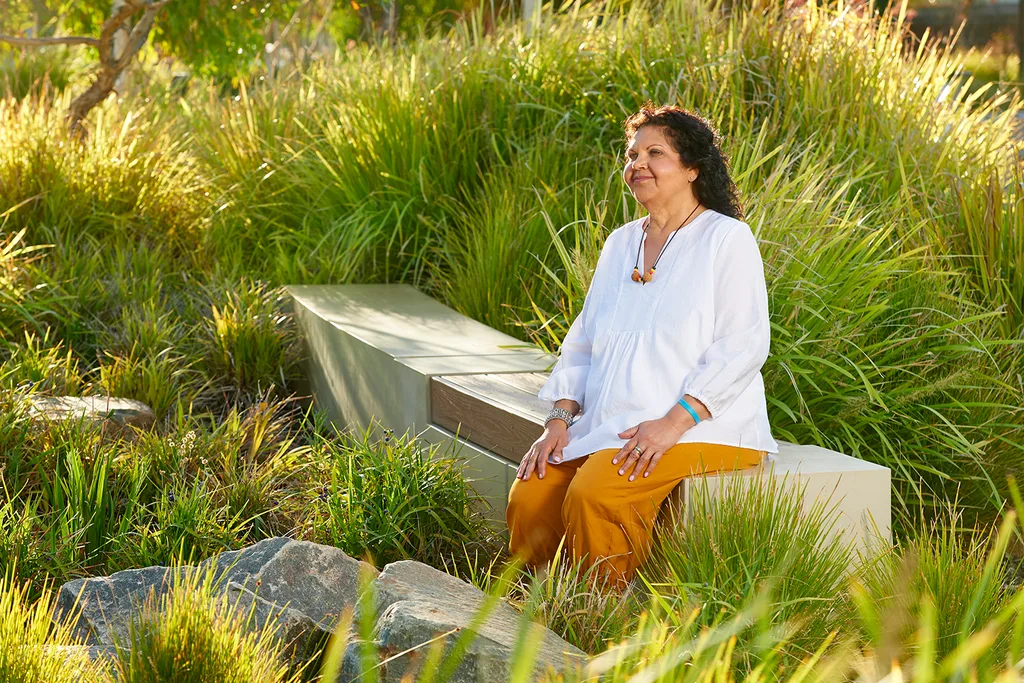 A woman in a white shirt sits on a bench surrounded by lush greenery.