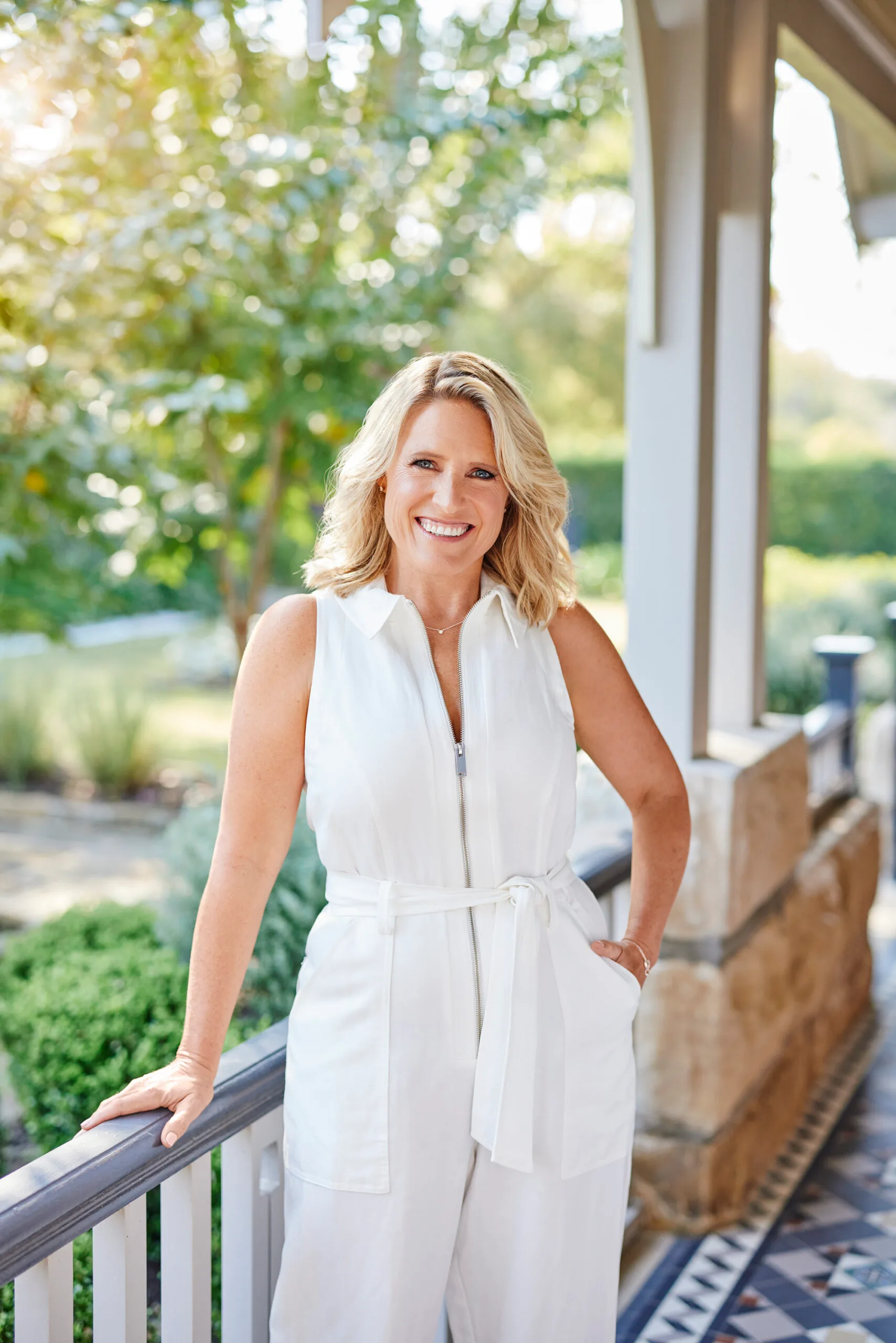 Smiling woman in a white jumpsuit standing on a porch with greenery in the background.
