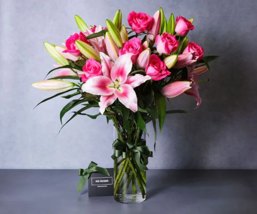 A bouquet of pink roses and lilies in a glass vase with a green ribbon, labeled "Mr Roses," on a gray background.