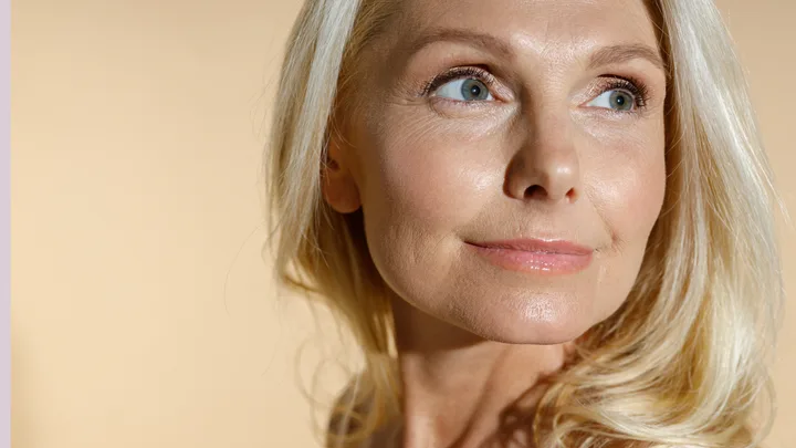 Portrait of a smiling woman with long blonde hair looking to the side against a beige background.