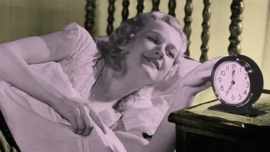 A woman in bed happily reaching for an alarm clock on a bedside table.