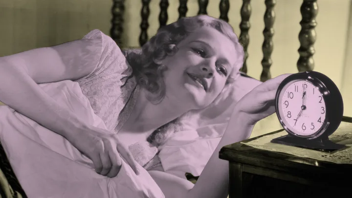 A woman in bed happily reaching for an alarm clock on a bedside table.