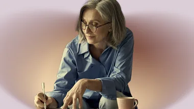 Elderly woman with glasses writing, sitting relaxed with a cup, in a blue shirt against a soft gradient background.