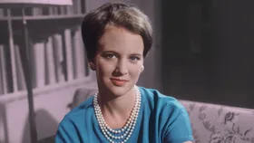 A woman in a blue dress with pearl necklace and earrings, sitting indoors with a bookshelf in the background.