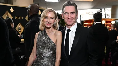 A woman in a silver dress and a man in a suit smile at an event with a champagne display in the background.