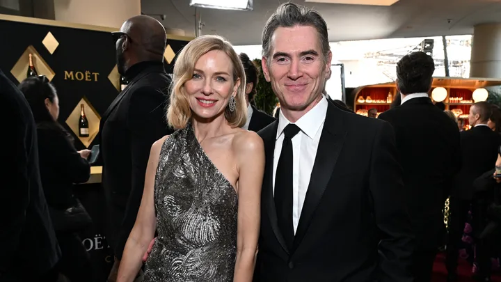 A woman in a silver dress and a man in a suit smile at an event with a champagne display in the background.