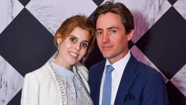 A smiling couple dressed formally poses in front of a patterned background.
