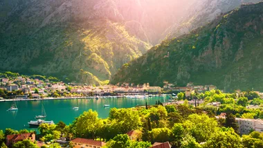 A scenic view of Kotor, Montenegro, showing the town, bay, and surrounding mountains under sunlight.
