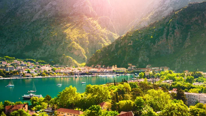 A scenic view of Kotor, Montenegro, showing the town, bay, and surrounding mountains under sunlight.
