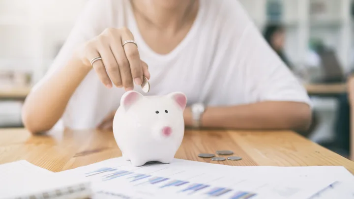 Person placing coin in white piggy bank on table with documents and coins.