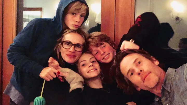 Family selfie with five people smiling indoors, one wearing glasses and another in a hoodie, wooden door in background.
